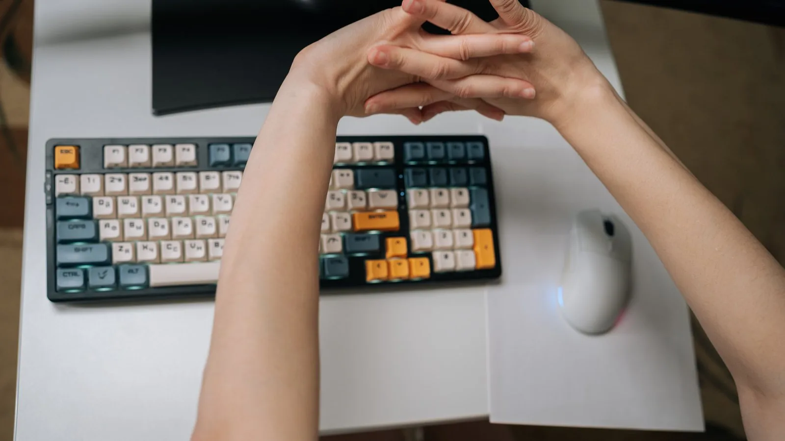 Two hands stretched and interlocked above a low, flat keyboard and mouse on a white desk
