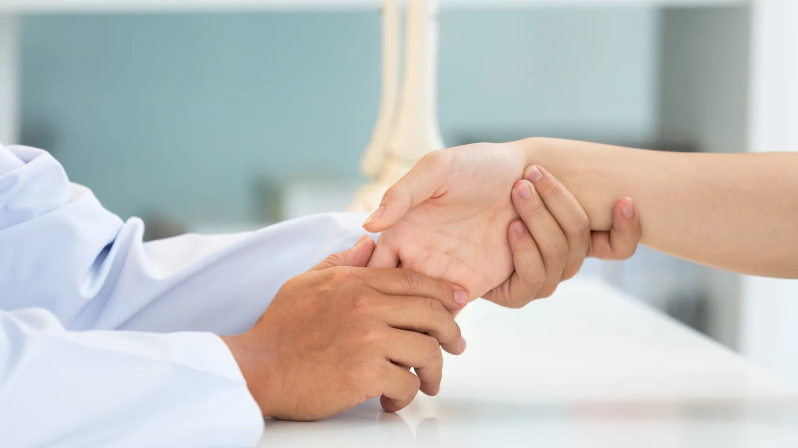 A clinician in a white coat examining a patient's wrist and hand across a consulting table