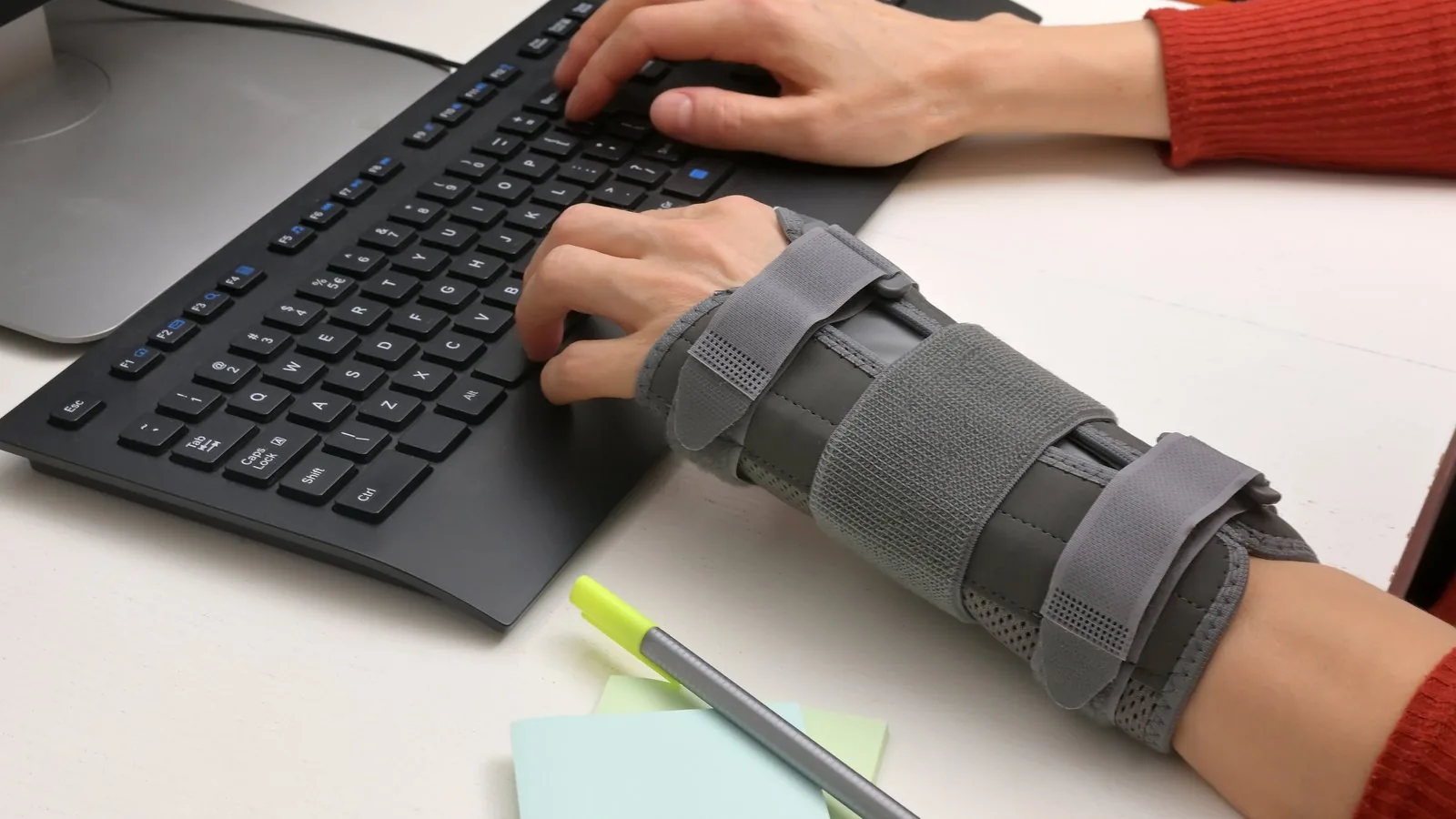 A hand typing on a keyboard while wearing a rigid grey wrist brace with adjustable straps
