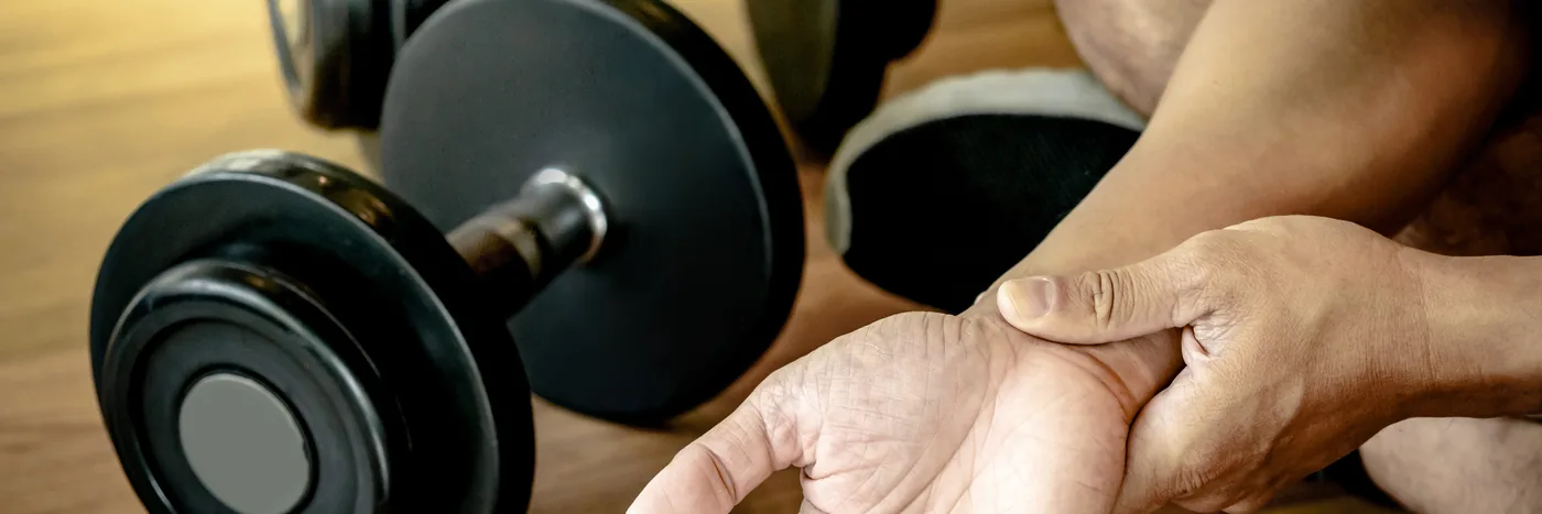 A person seated beside a dumbbell on a gym floor, holding their wrist with the opposite hand.