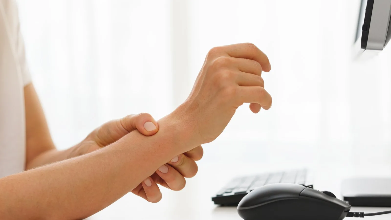 A pair of hands at a desk, one hand gripping the opposite wrist beside a keyboard and mouse