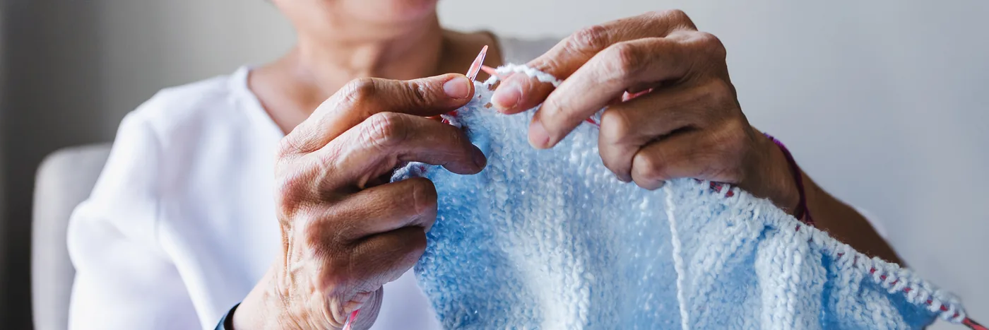 The hands of an older woman knitting a pale blue garment on circular needles.