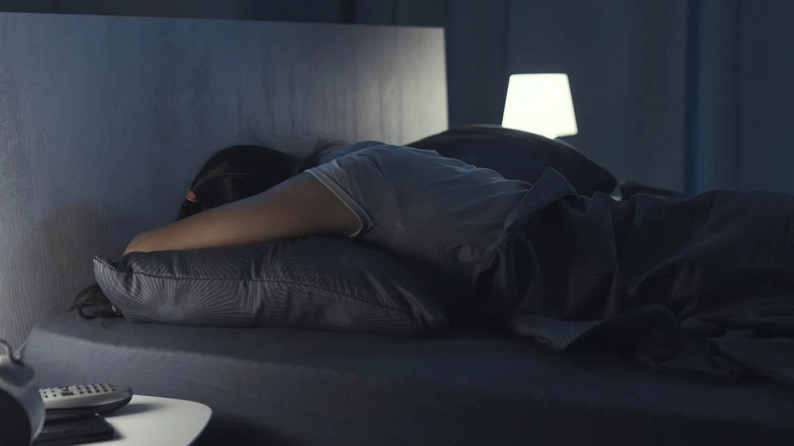 A person asleep on their side in a dark bedroom lit by a single bedside lamp