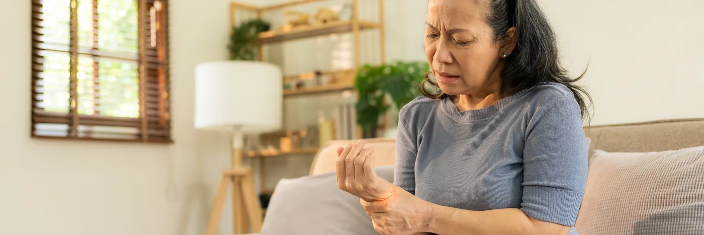 An older woman on a sofa at home wincing as she works the fingers of one hand with the other.